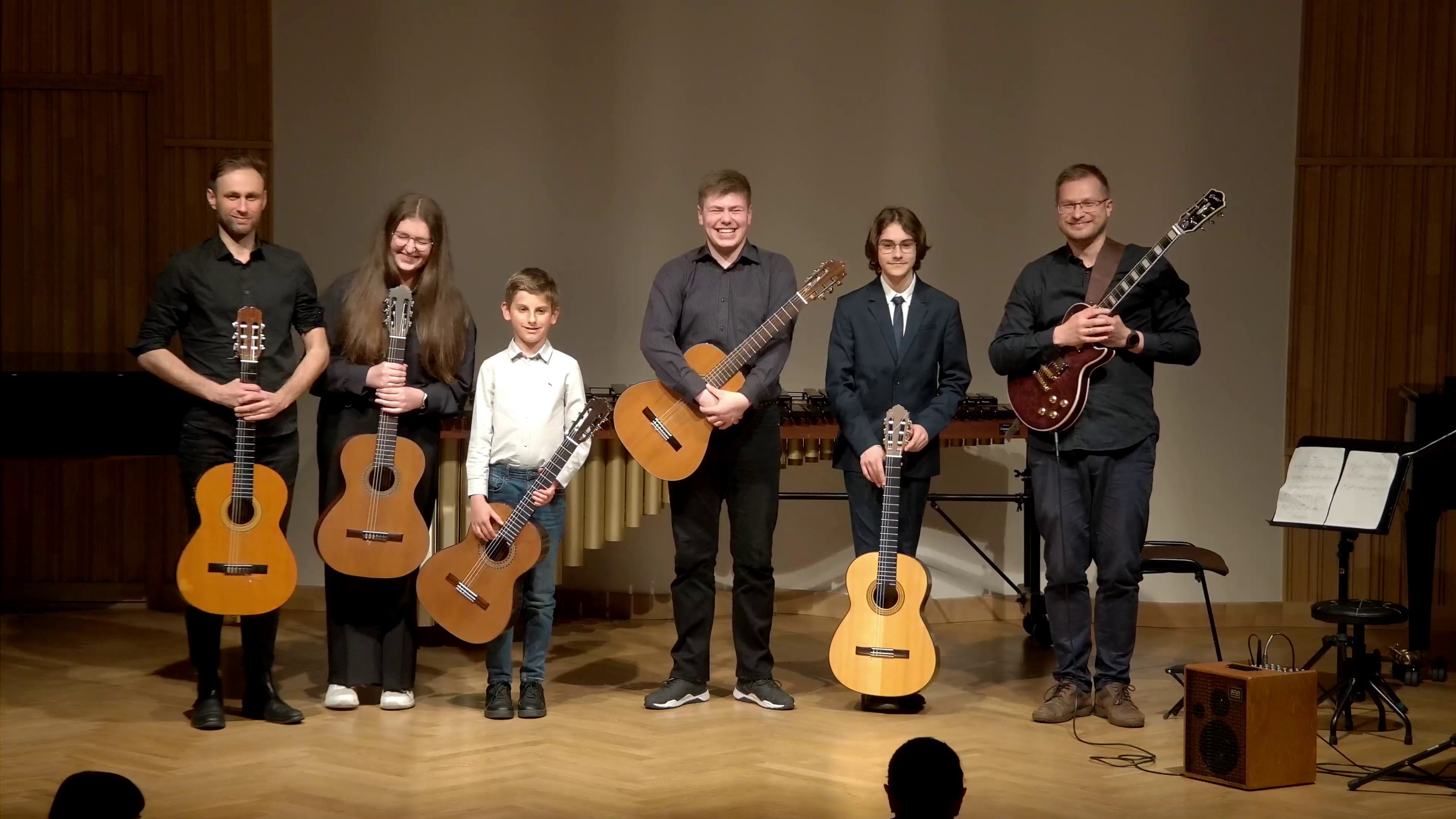 Zdjęcie. Na estradzie sali koncertowej stoją gitarzyści - uczniowie i nauczyciele - wykonawcy koncertu gitarowego Wszyscy trzymają gitary i się uśmiechają.