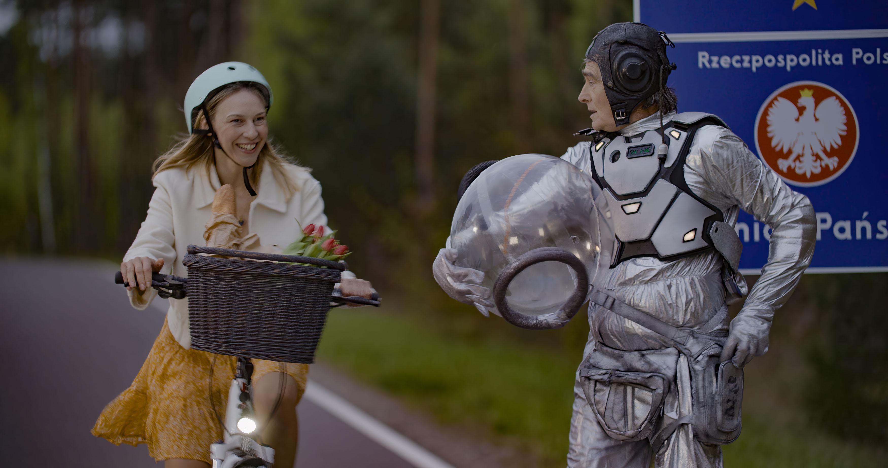 Mężczyzna w stroju astronauty stoi na tle znaku granicy Polski, obok przejeżdża uśmiechnięta młoda kobieta na rowerze