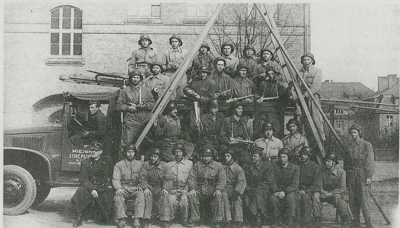 Zdjęcie przedstawia grupę mężczyzn, strażaków, ustawionych do zdjęcia grupowego na tle wozu strażackiego, drabiny pożarnicze ustawione są na kształt odwróconej litery V