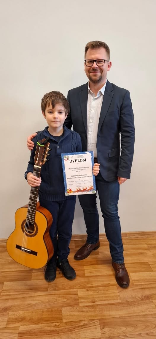 Na zdjęciu widoczny jest uczeń wraz ze swoim nauczycielem. Uczeń trzyma w dłoniach gitarę oraz dyplom.