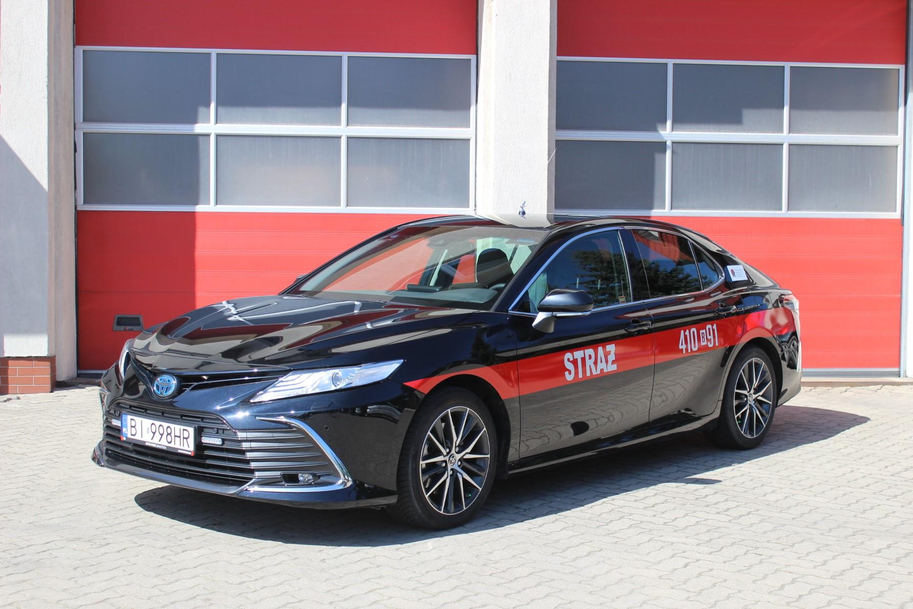 Zdjęcie przedstawia samochód operacyjny TOYOTA Camry stojący na tle strażnicy.