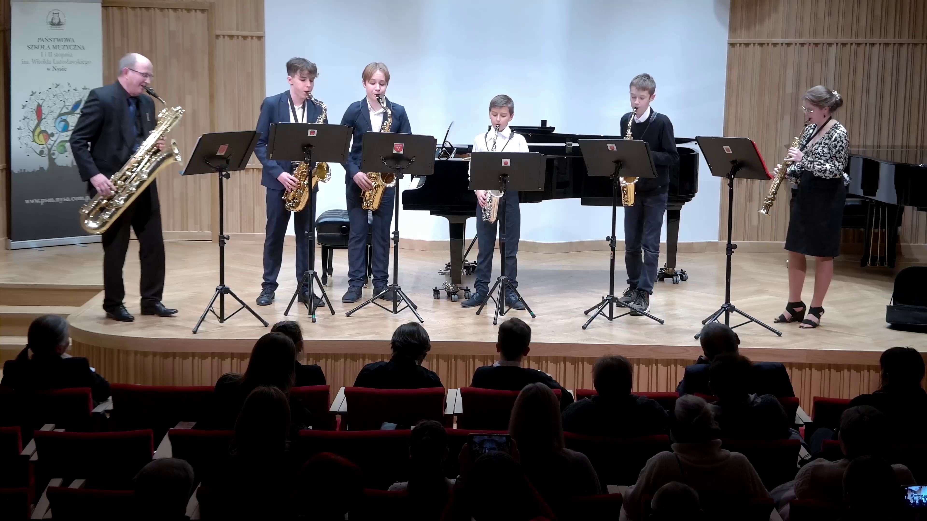 Zdjęcie. Na estradzie Sali Koncertowej PSM I i II stopnia w Nysie występuje zespół saksofonowy, pięciu uczniów z nauczycielem. Przed wykonawcami stoją pulpity z nutami. W dolnej części zdjęcia widać publiczność.