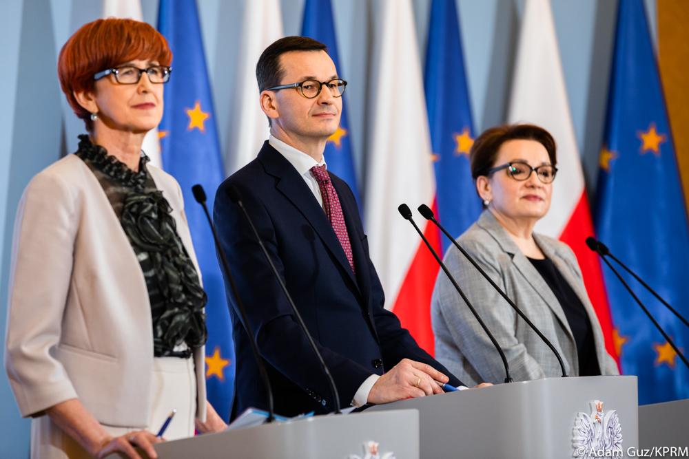 Od lewej: minister Elżbieta Rafalska, premier Mateusz Morawiecki oraz minister Anna Zalewska stoją podczas konferencji prasowej na tle biało-czerwonych flag i flag Unii Europejskiej.