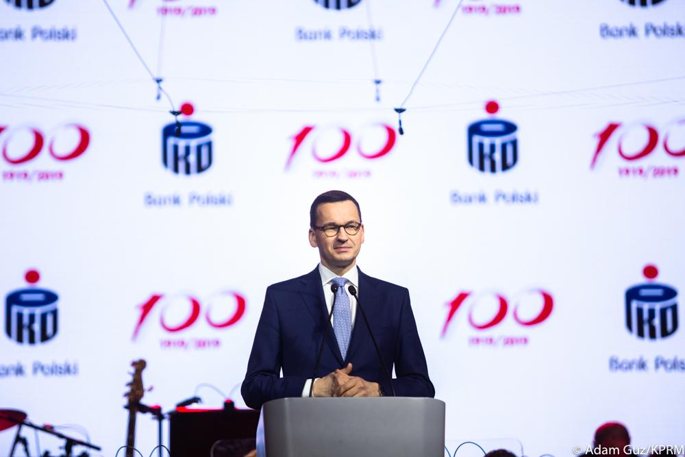 Premier Mateusz Morawiecki stoi przy mównicy na tle logotypów PKO BP.