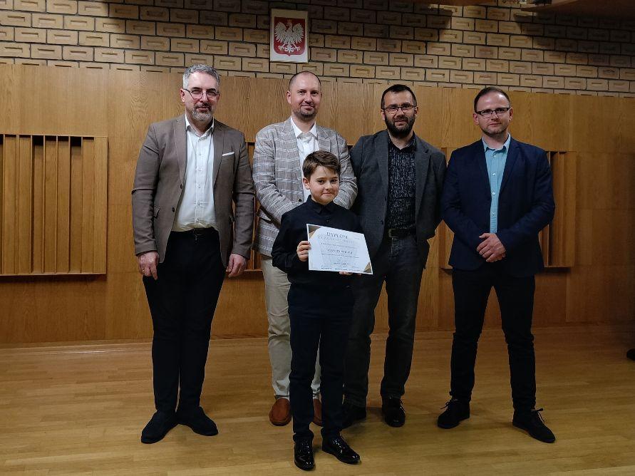 Z przodu uczeń trzymający dyplom laureata, za nim czterech członków Jury. W tle, po środku widoczne godło Polski.