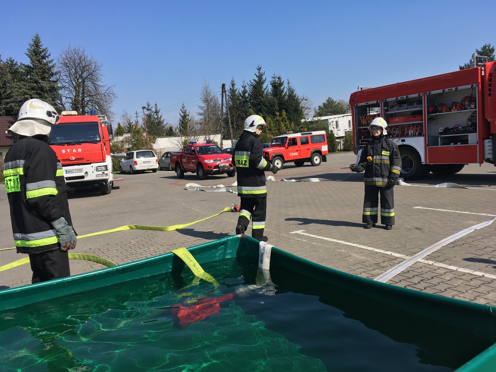 Zdjęcie przedstawia trzech strażaków OSP podczas wypompowywania wody ze zbiornika brezentowego, w w tle samochody pożarnicze.