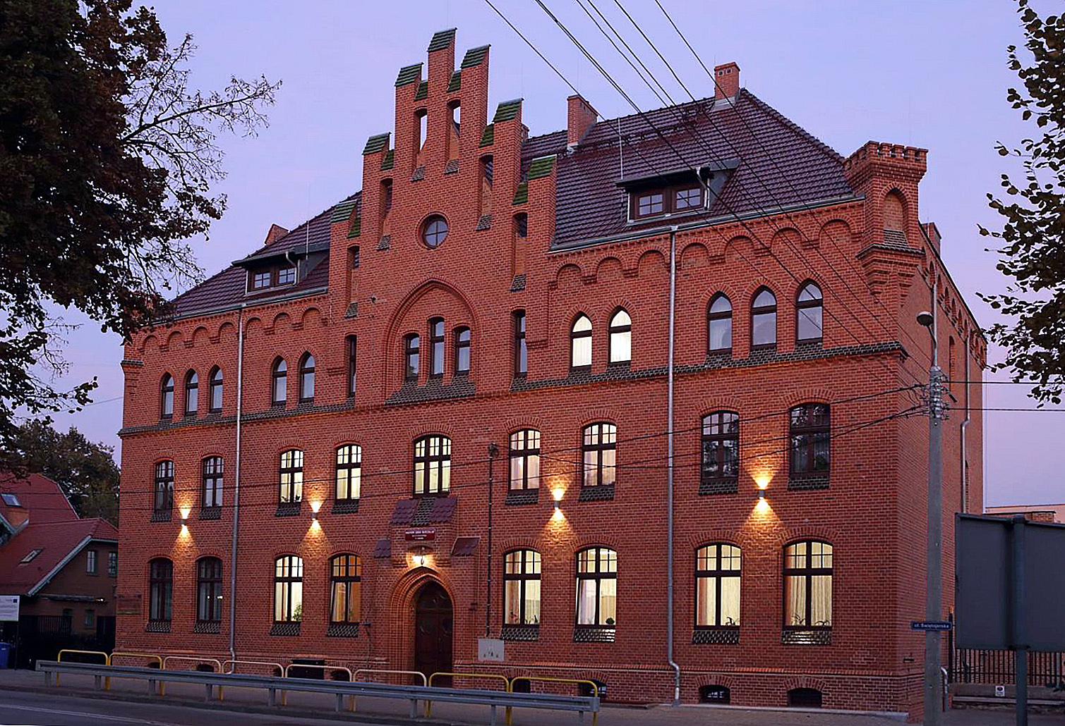 Kolorowa fotografia przedstawiająca gmach szkoły muzycznej. Budynek 3 kondygnacyjny. Zdjęcie wykonane późnym popołudniem lub wieczorem. Widoczne oświetlenie wewnętrzne w niektórych pomieszczeniach