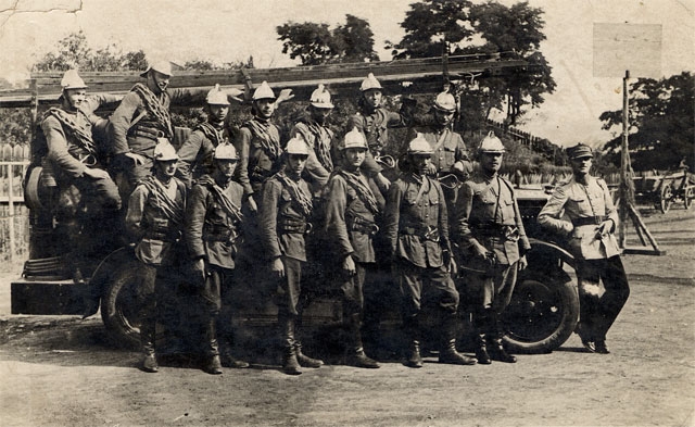 Załoga (14 strażaków) Miejskiej Zawodowej Straży Pożarnej w Nowym Sączu na placu strażnicy przy ul. Grybowskiej przy bramie wjazdowej w 1930 roku. W tle widać fragment placu targowego. 