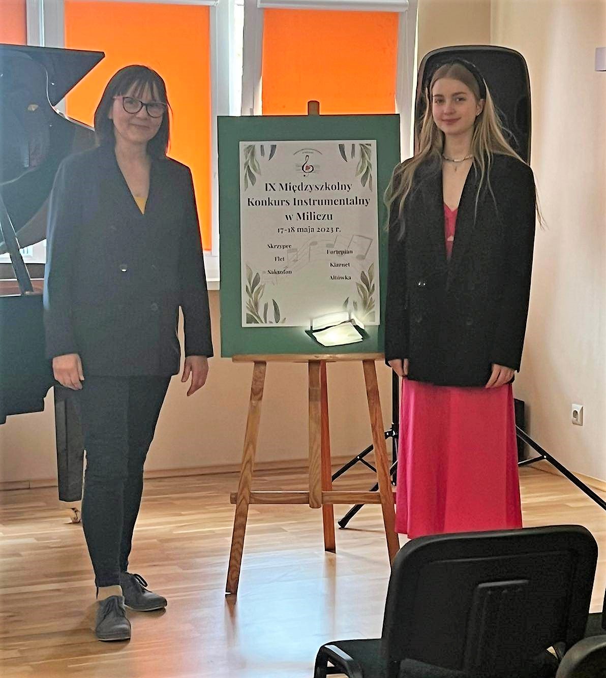 dwie osoby stojące na scenie obok fortepianu, między nimi sztaluga z plakatem konkursu instrumentalnego
