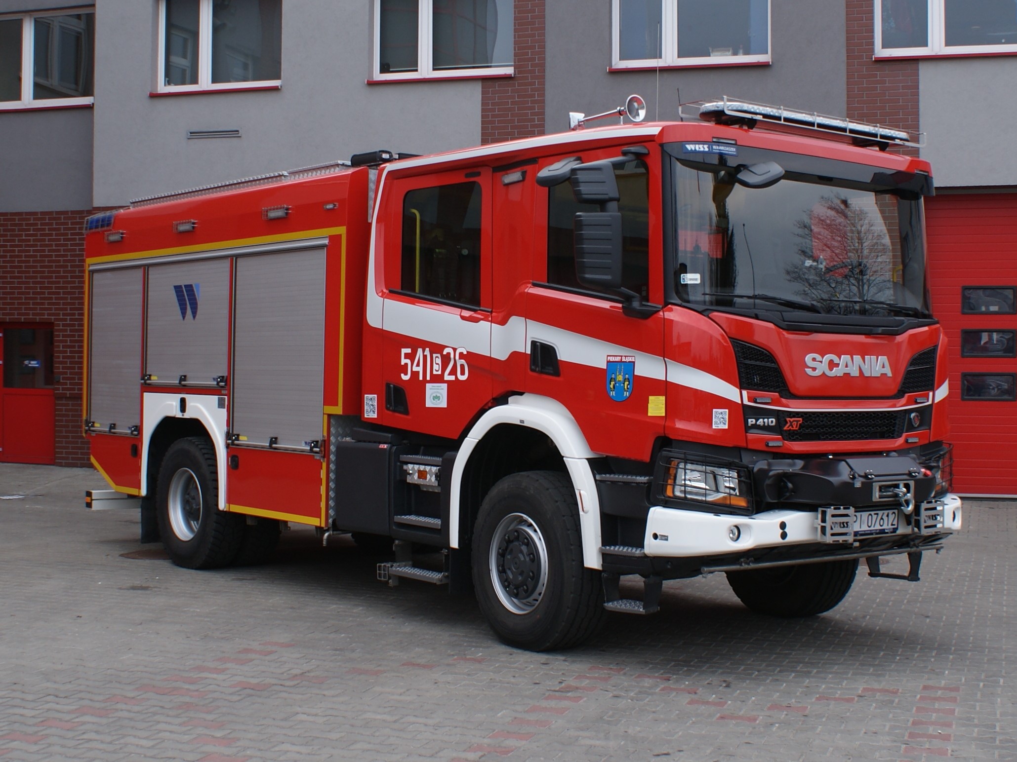Zdjęcie przedstawia ciężki samochód ratowniczo-gaśniczy marki Scania w kolorze czerwonym ma budynku komendy
