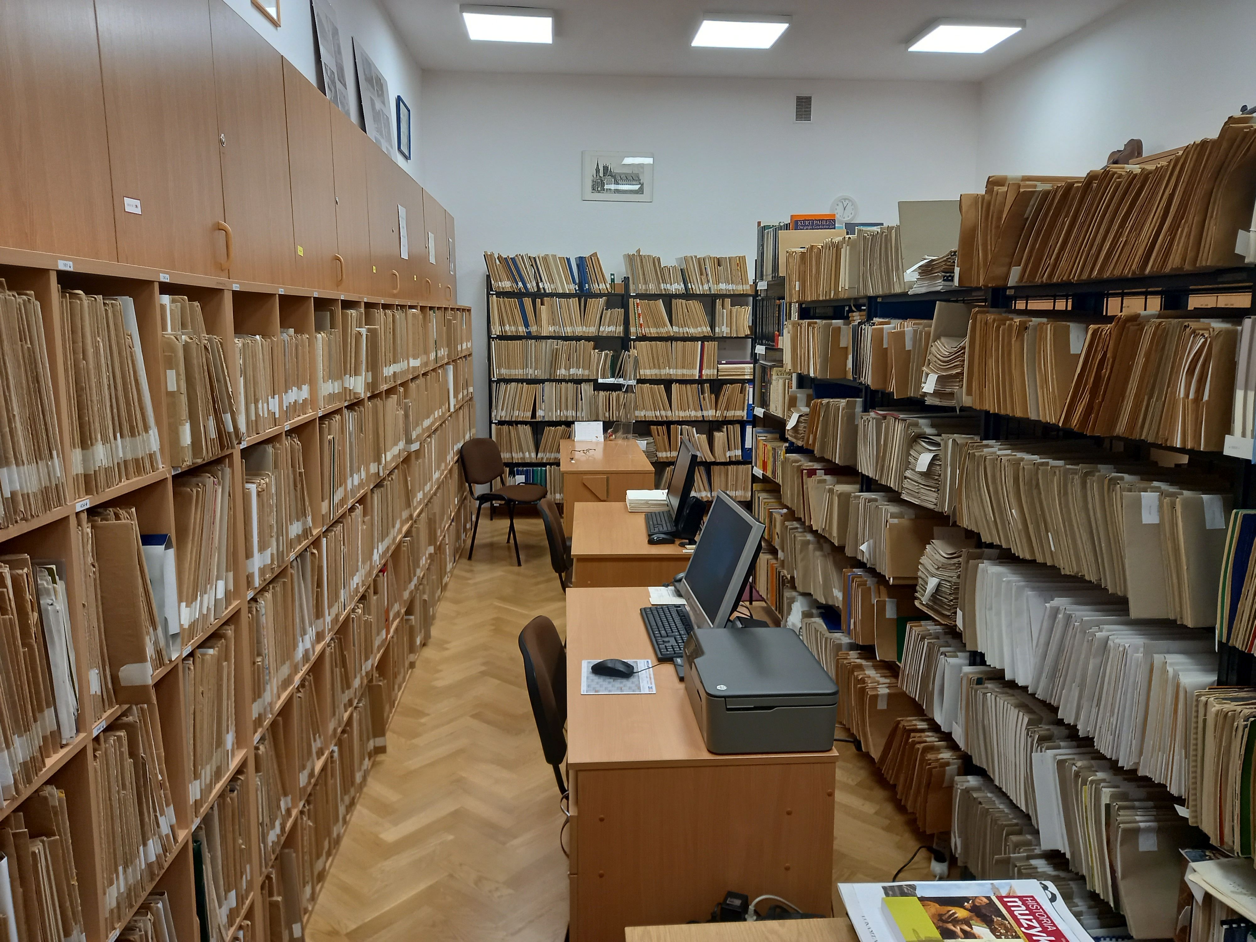 Zdjęcie. W środku biurka ze stanowiskami komputerowymi. Z lewej i prawej strony regały z książkami, nutami oraz szafki z płytami. W tle również regały z nutami.