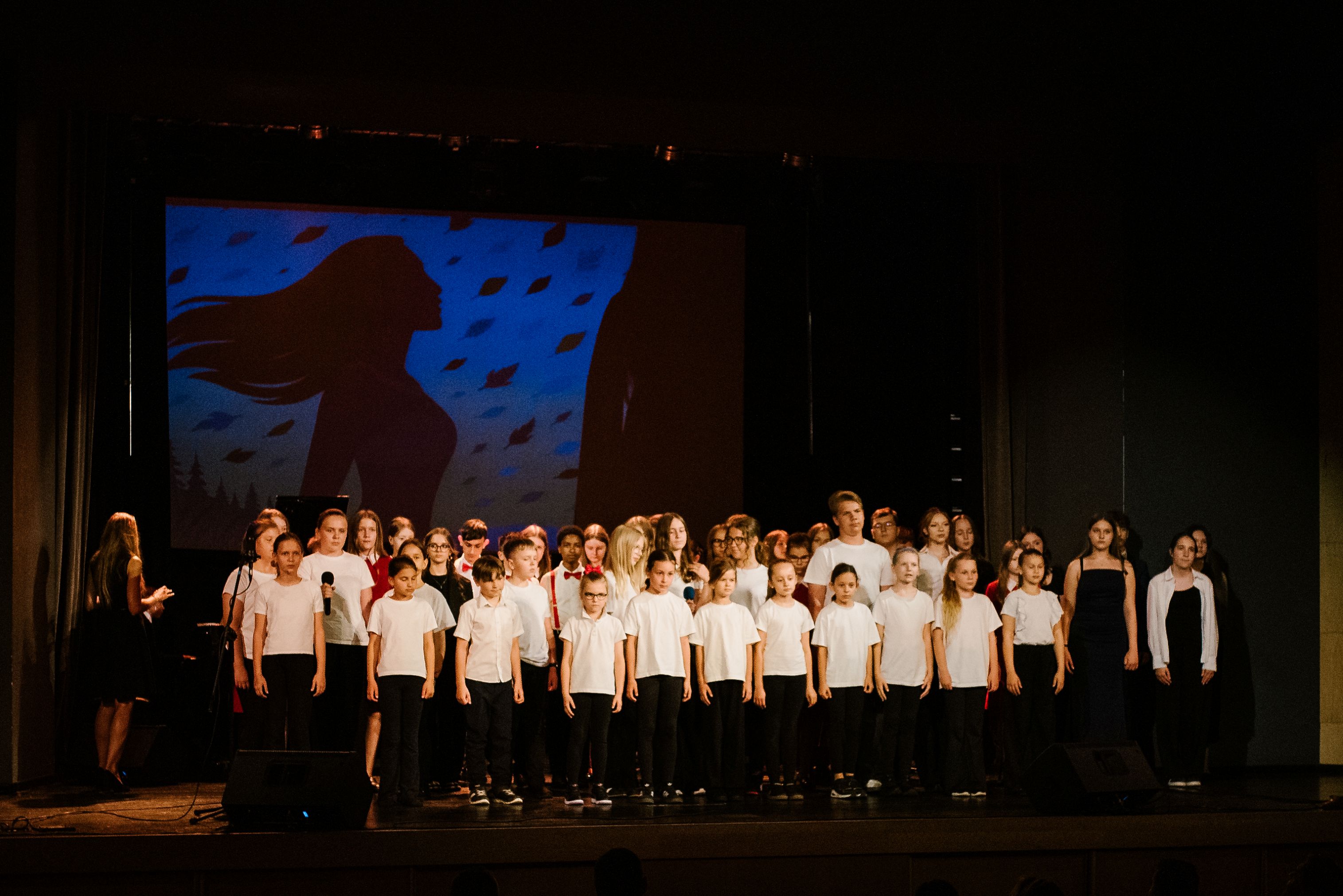 Na zdjęciu widoczna jest duża grupa dzieci ustawiona w kilku rzędach na scenie.