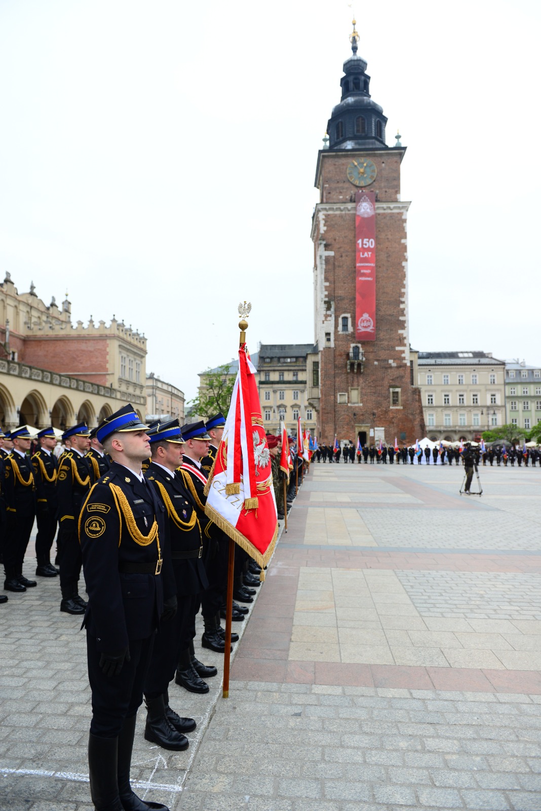 Na Rynku Głównym Krakowa stoją pododdziały z sztandarami, w tle wieża ratuszowa 