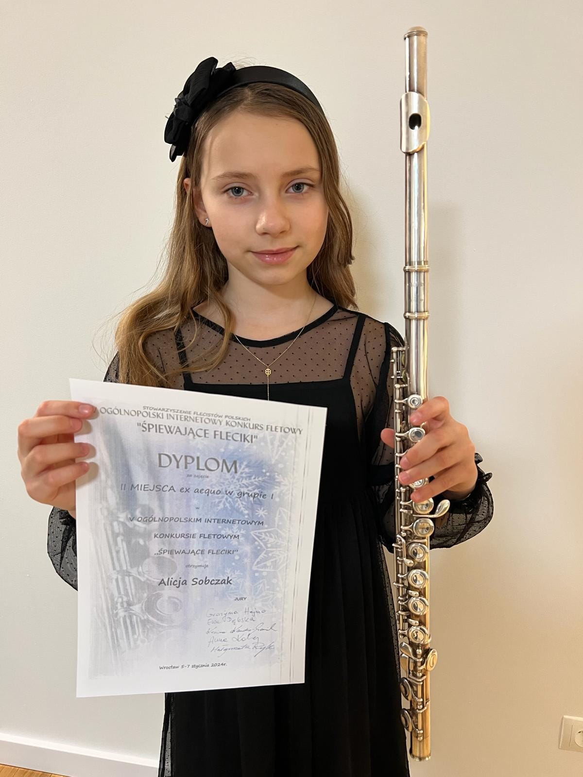 Na zdjęciu widoczna uśmiechnięta dziewczynka w czarnej sukience, na głowie na czarną opaskę. W rękach trzyma flet oraz dyplom konkursowy. 