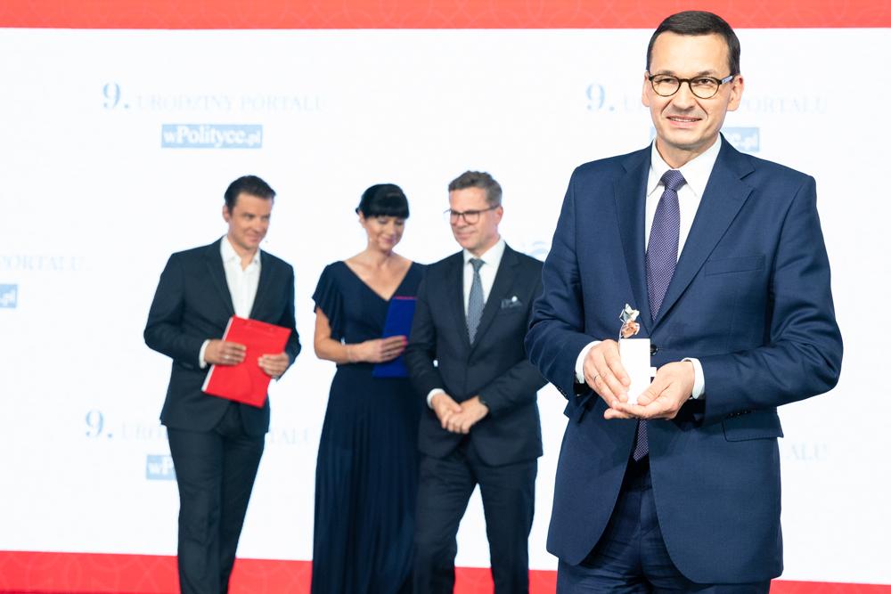 Premier Mateusz Morawiecki ze statuetką w ręce, a w tle stoją prowadzący galę.
