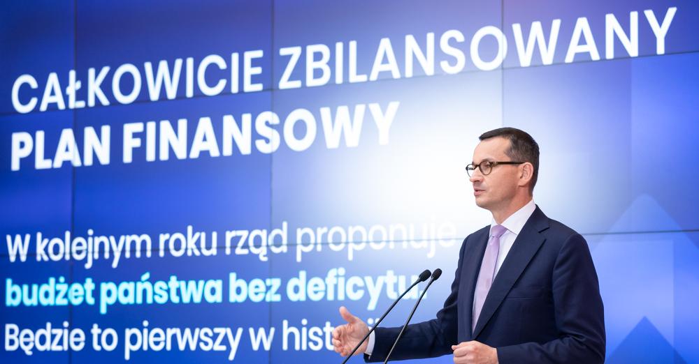 Premier przy mównicy, a w tle prezentacja.