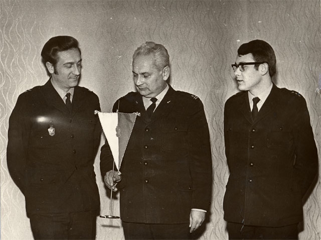 Stoją od lewej w mundurach bez czapek widoczni do pasa: ppor.poż. Bienias Antoni, mjr poż Strojny Józef (który trzyma w ręku proporczyk) oraz por. poż. Raczek Zbigniew