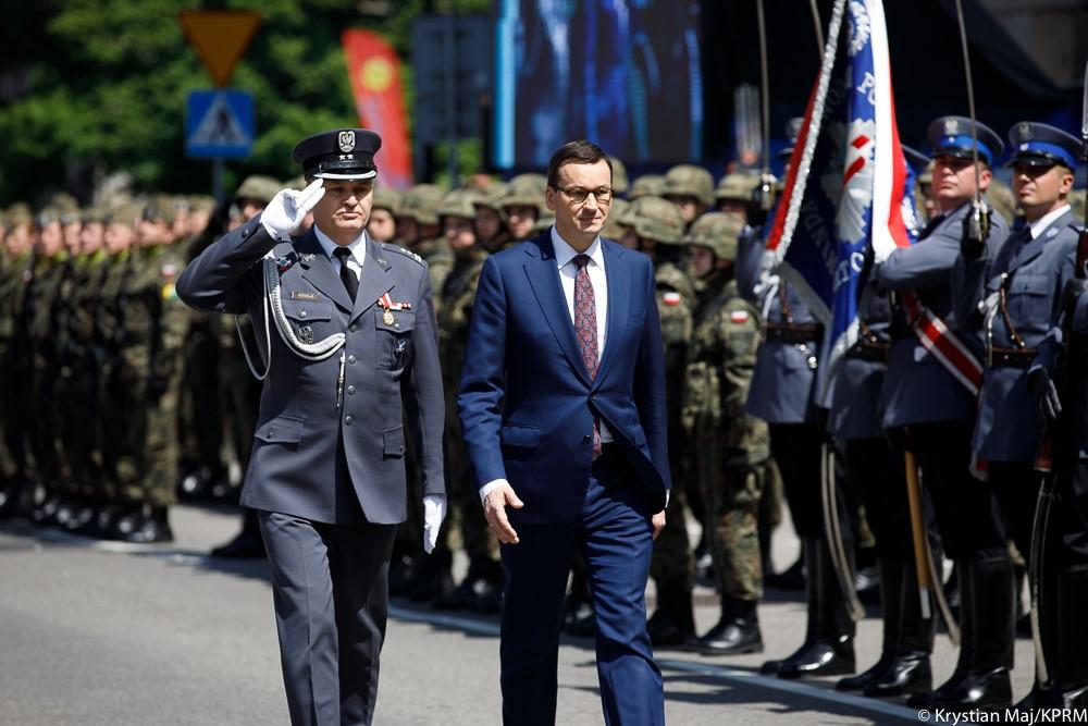 Premier Mateusz Morawiecki na tle sztandarów i wojska.