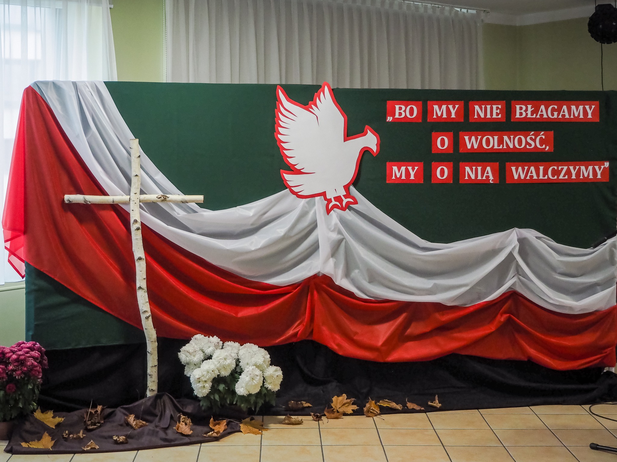 Na fotografii patriotyczna scenografia z napisem „Bo my nie błagamy o wolność, my o nią walczymy”, orzeł z biało- czerwoną, krzyż brzozowy i białe chryzantemy.