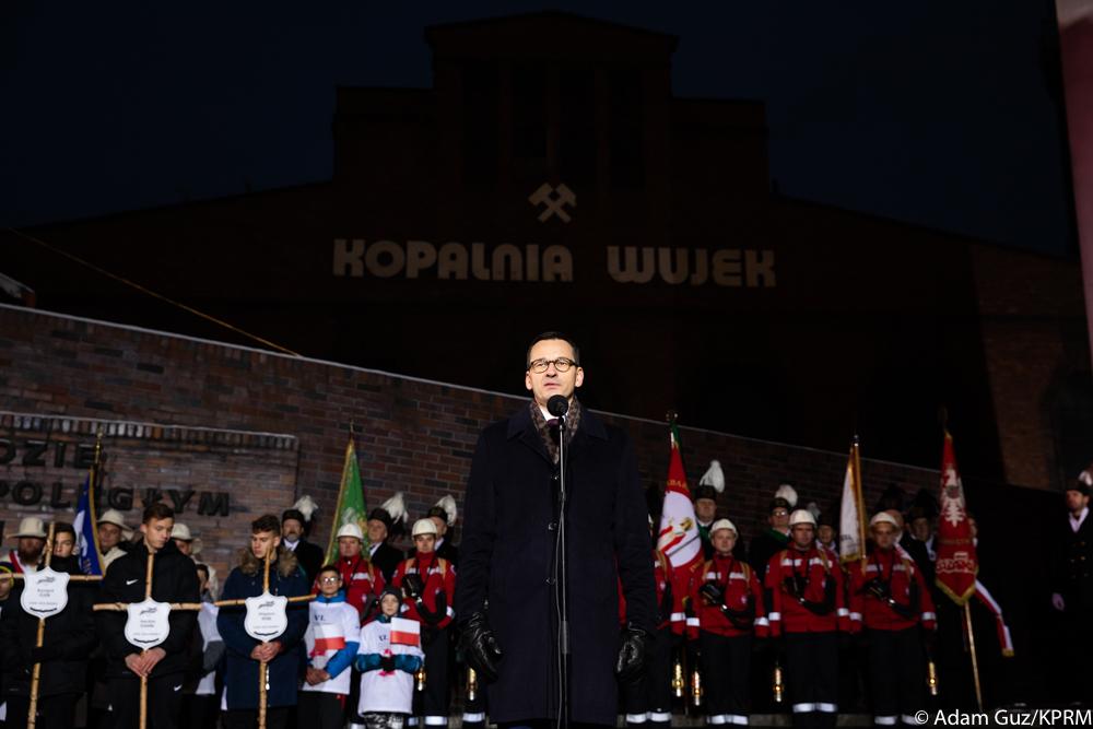 Premier Mateusz Morawiecki przemawia przed mikrofonem, w tle górnicy.