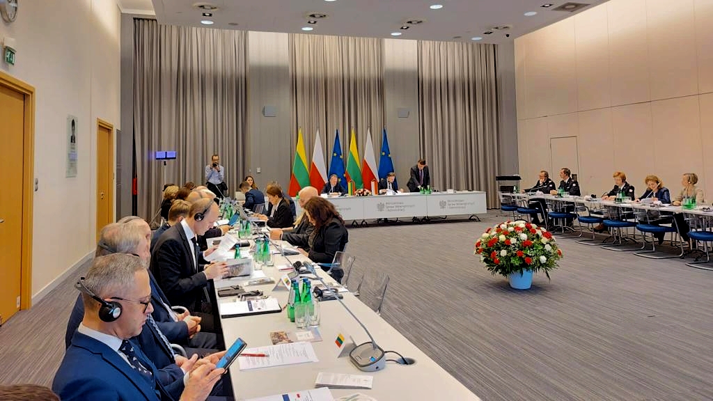 Stół konferencyjny z siedzącymi przy nim uczestnikami spotkania w MSWiA. Stoły ustawione w kształcie litery U na środku sali stoi wazon z kwiatami. Za stołem prezydialnym widoczne flagi Litwy, Polski i Unii Europejskiej. 
