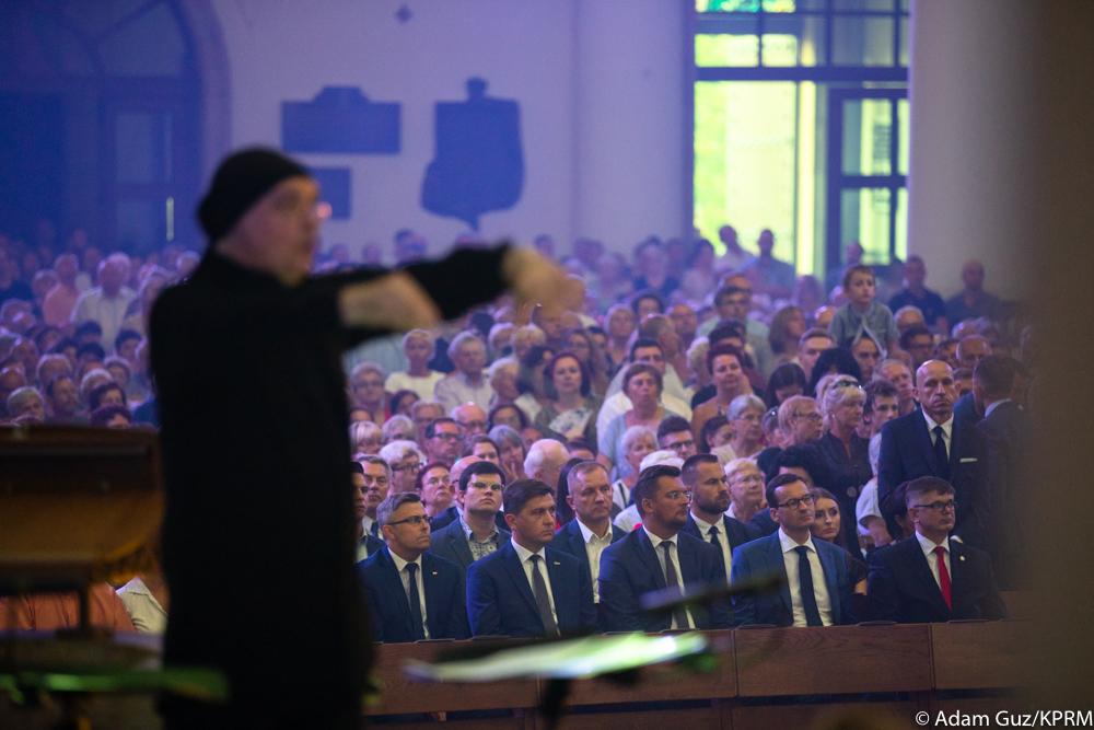 Dyrygent, a w tle premier Mateusz Morawiecki i uczesnicy uroczystości oglądają koncert.