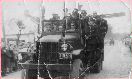 Zdjęcie historyczne KP PSP w Kozienicach przedstawiający wóz strażacki przejeżdżający przez ulicę miasta. Na chodnikach zgromadzeni ludzie odwróceni w stronę wozu. W wozie siedzą strażacy lub stoją trzymając się karoserii.