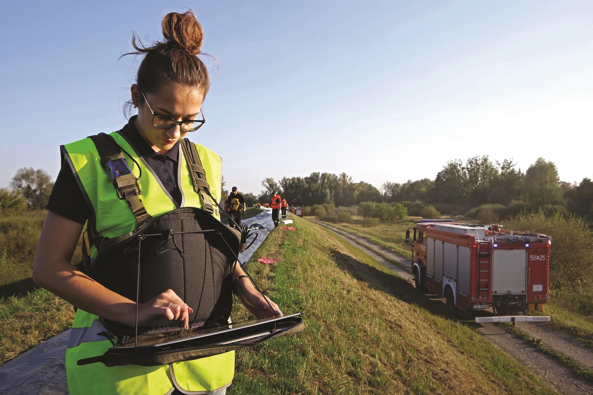A woman in a yellow reflective vest is standing with a laptop by the embankment of the river, below the fire station