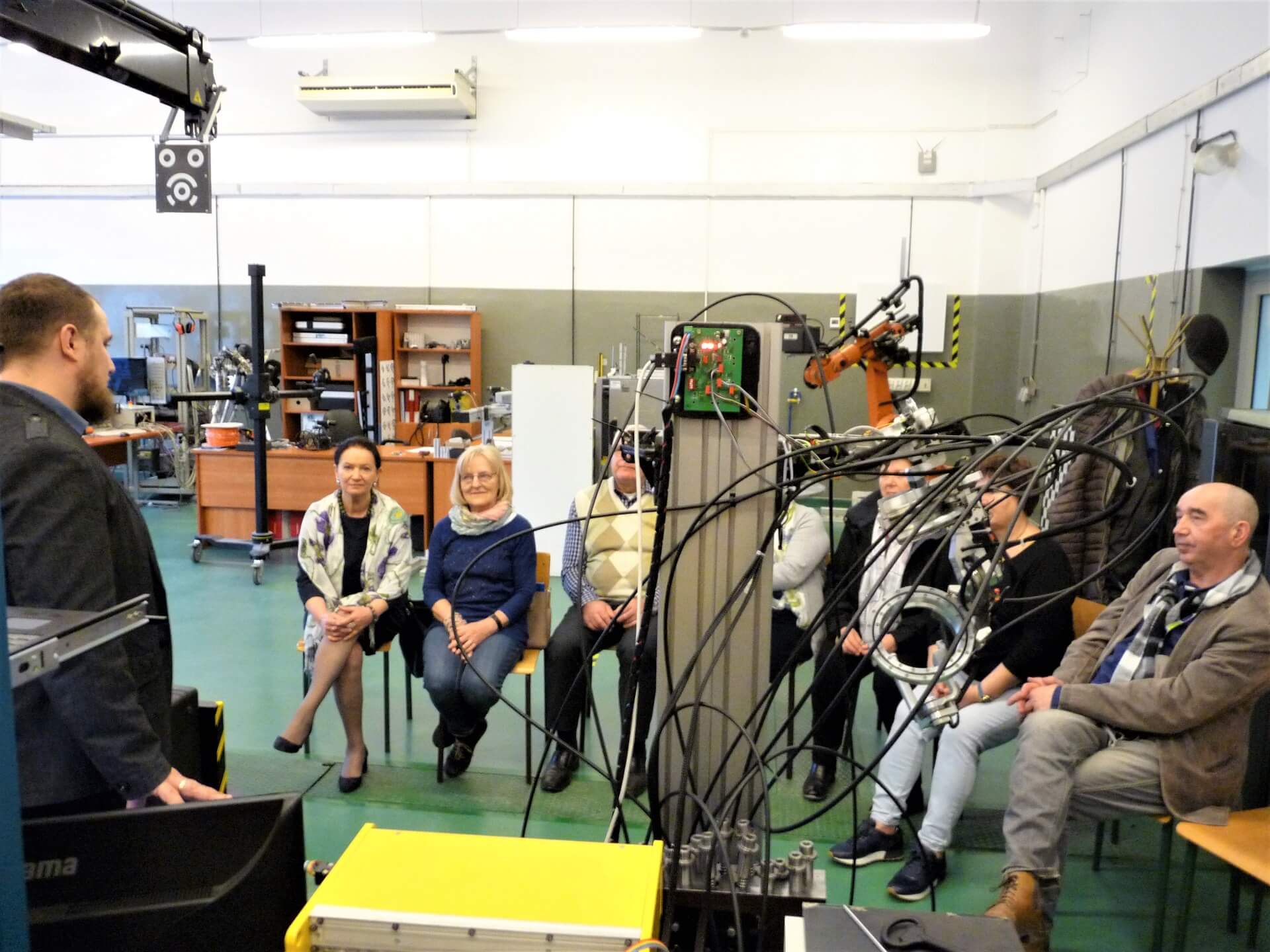 Zajęcia na hali technologicznej, grupa seniorów siedzi w kręgu, słuchając prowadzącego. Na pierwszym planie specjalistyczne przyrządy