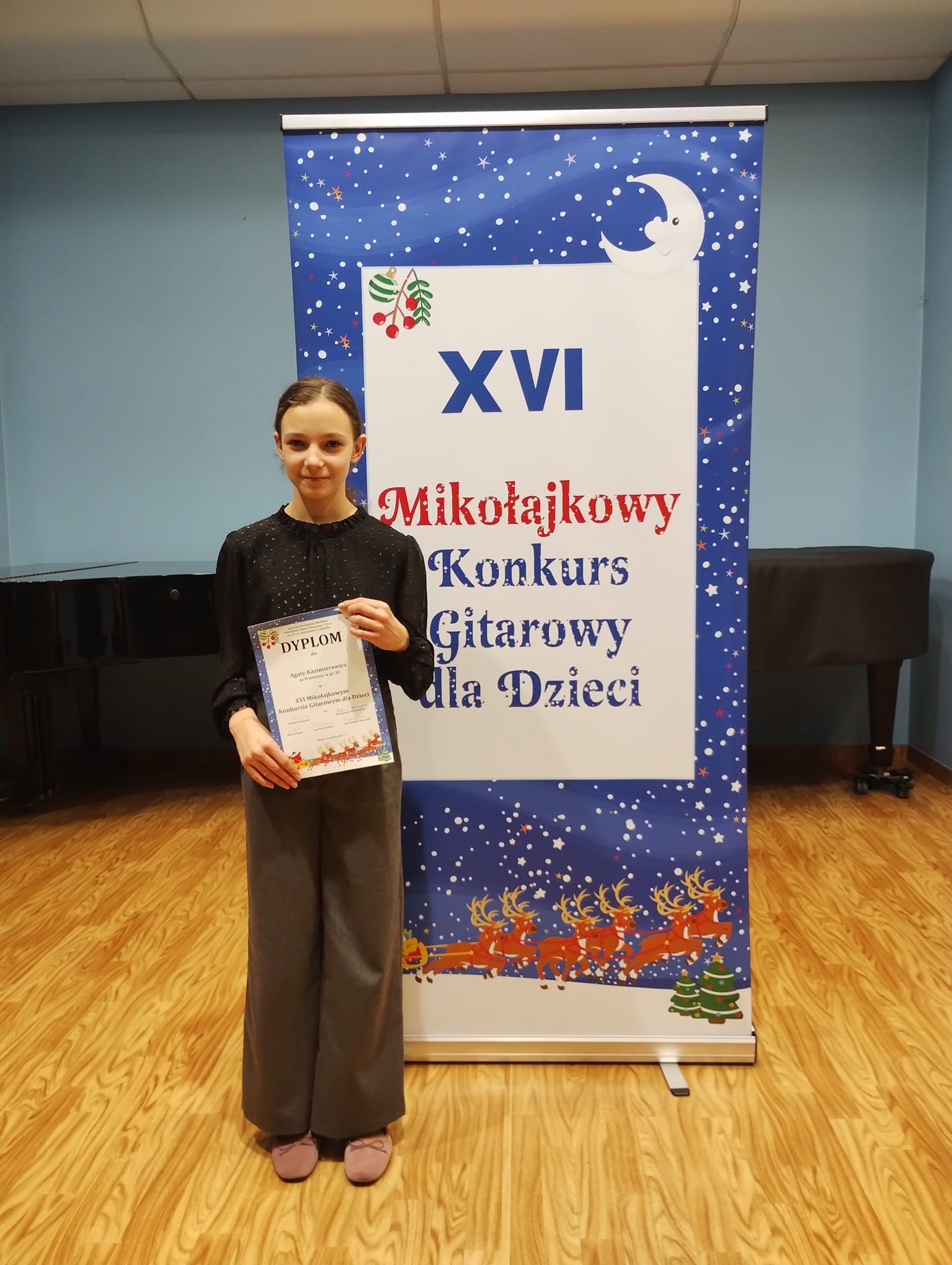 Na fotografii znajduje się uczennica Agata Kostanowicz trzymająca w rękach dyplom. Stoi na tle pionowego baneru konkursu. 
