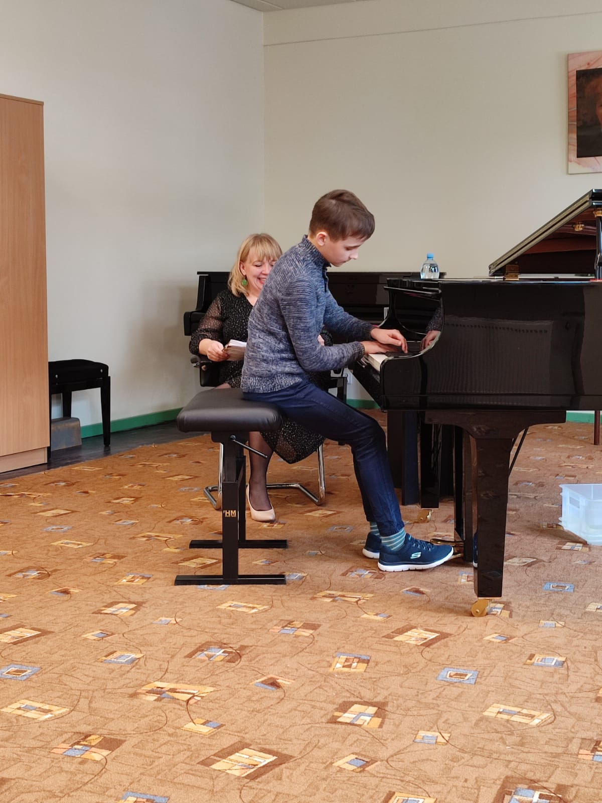 Uczeń siedzący przy fortepianie, obok niego nauczycielka. Po lewej stronie widoczna szafa. Na ścianie wisi portret Ignacego Jana Paderewskiego.