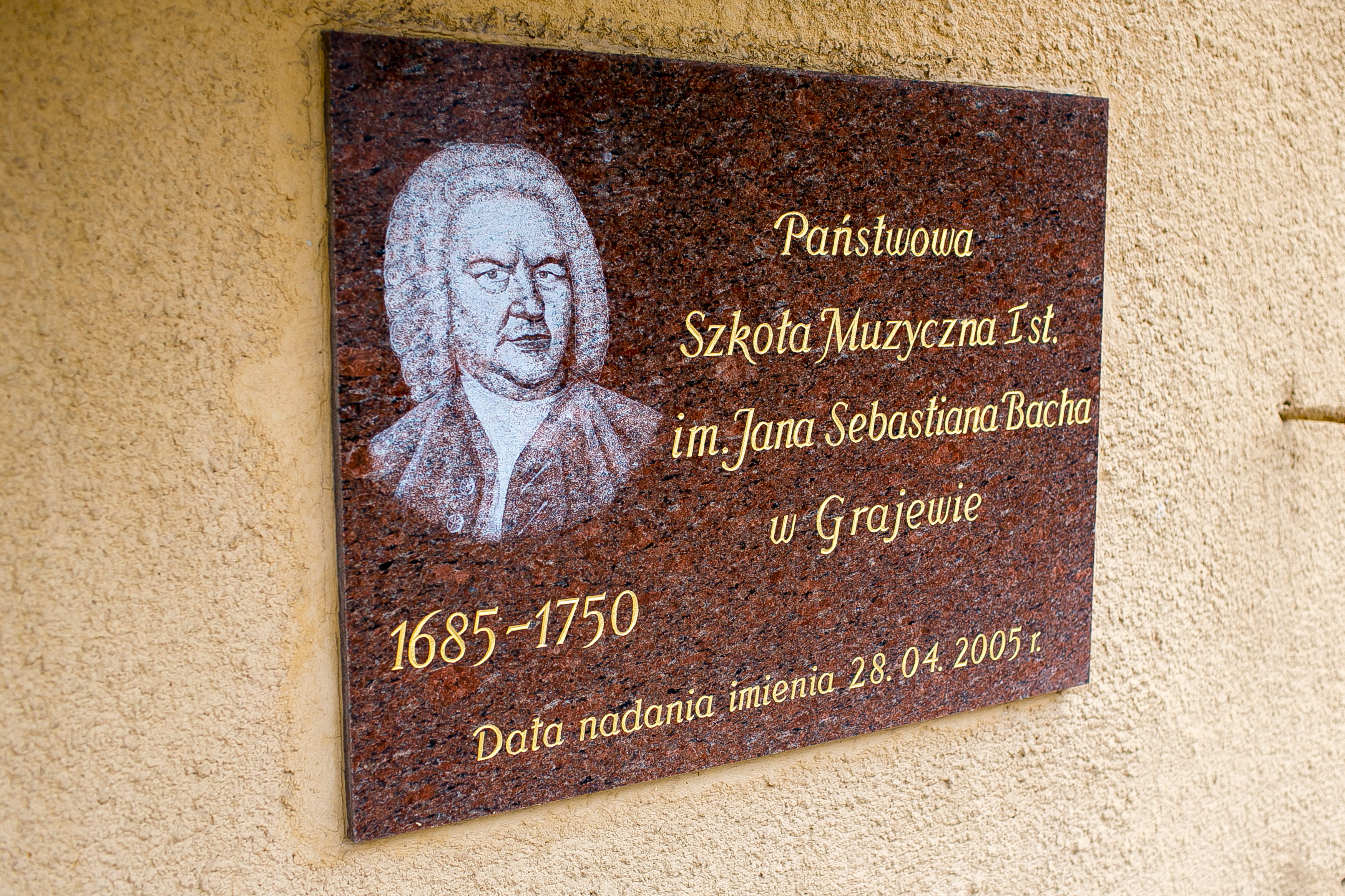 Marmurowa tablica przedstawiająca wizerunek kompozytora Jana Sebastiana Bacha z data nadania imienia szkole oraz tekstem "Państwowa Szkoła Muzyczna I st. im. Jana Sebastiana Bacha w Grajewie"