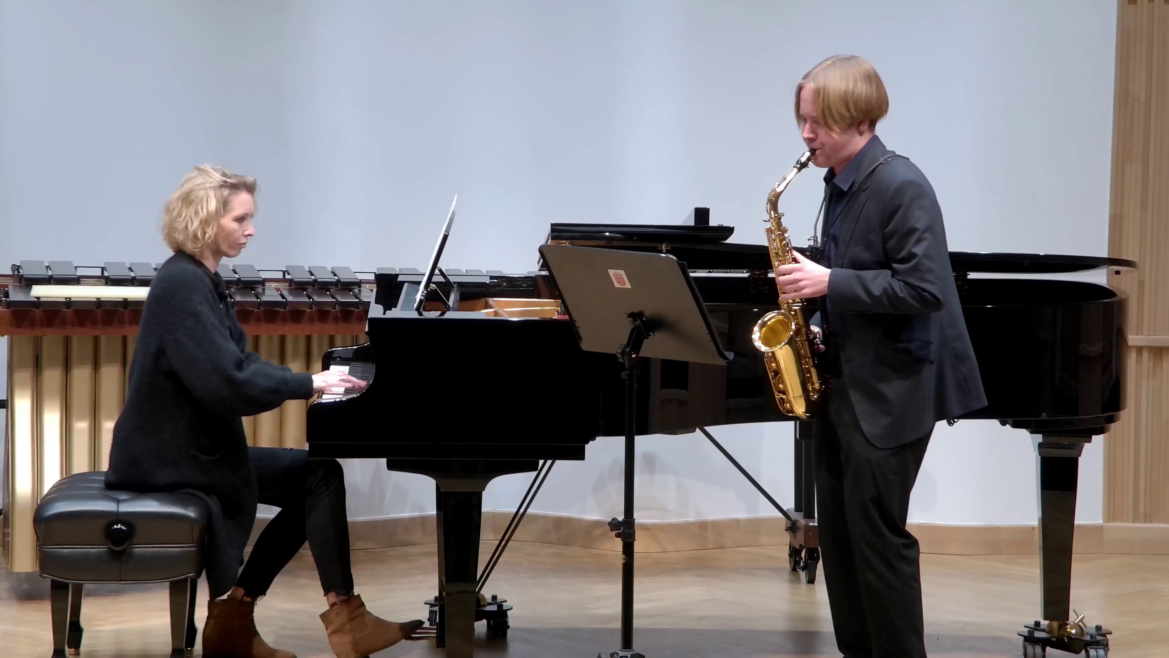 Zdjęcie. Na estradzie sali koncertowej występuje saksofonista z towarzyszeniem pianistki. Za fortepianem w tle stoi instrument perkusyjny - marimba.