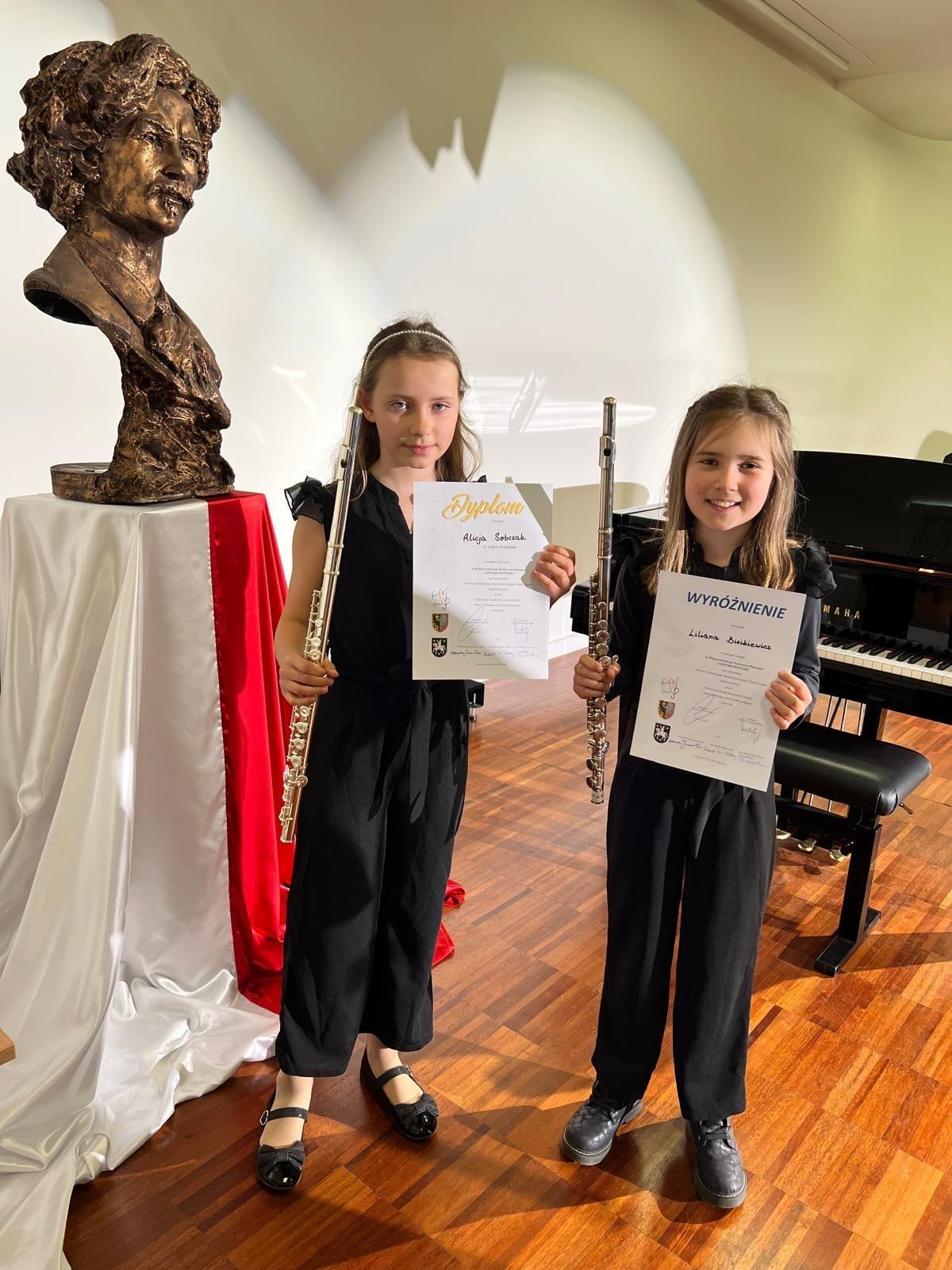 Na zdjęciu widnieją dwie uśmiechnięte dziewczynki, w jednej ręce trzymają dyplomy konkursowe, w drugiej flety poprzeczne. W tle znajduje się popiersie patrona szkoły z flagą Polski oraz czarny fortepian. 