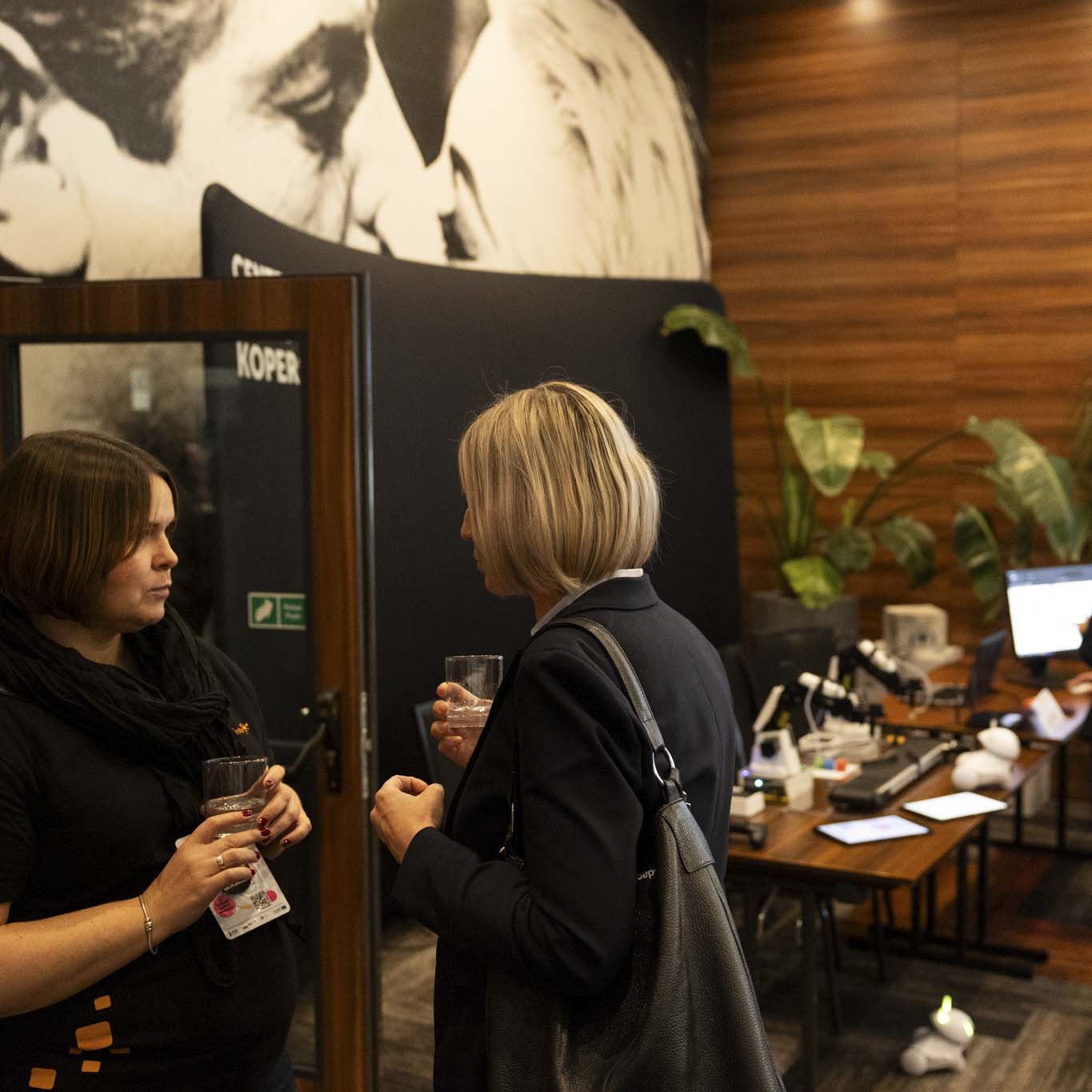 Dwie kobiety rozmawiają, w tle trzy osoby stoją i zainteresowaniem patrzą w ekran.