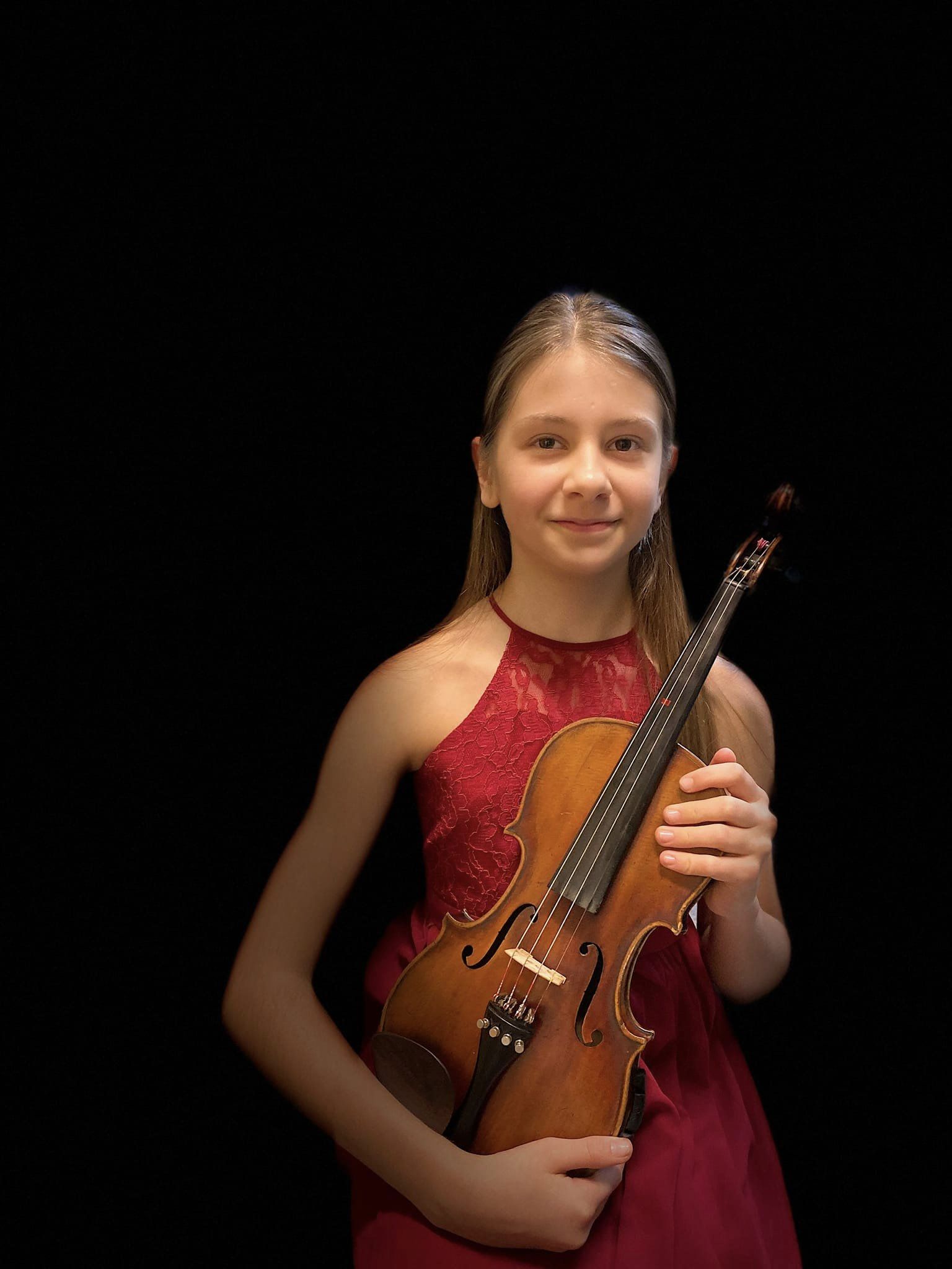 Na zdjęciu znajduje się uśmiechnięta dziewczynka w czerwonej sukience, w rękach trzyma skrzypce. 