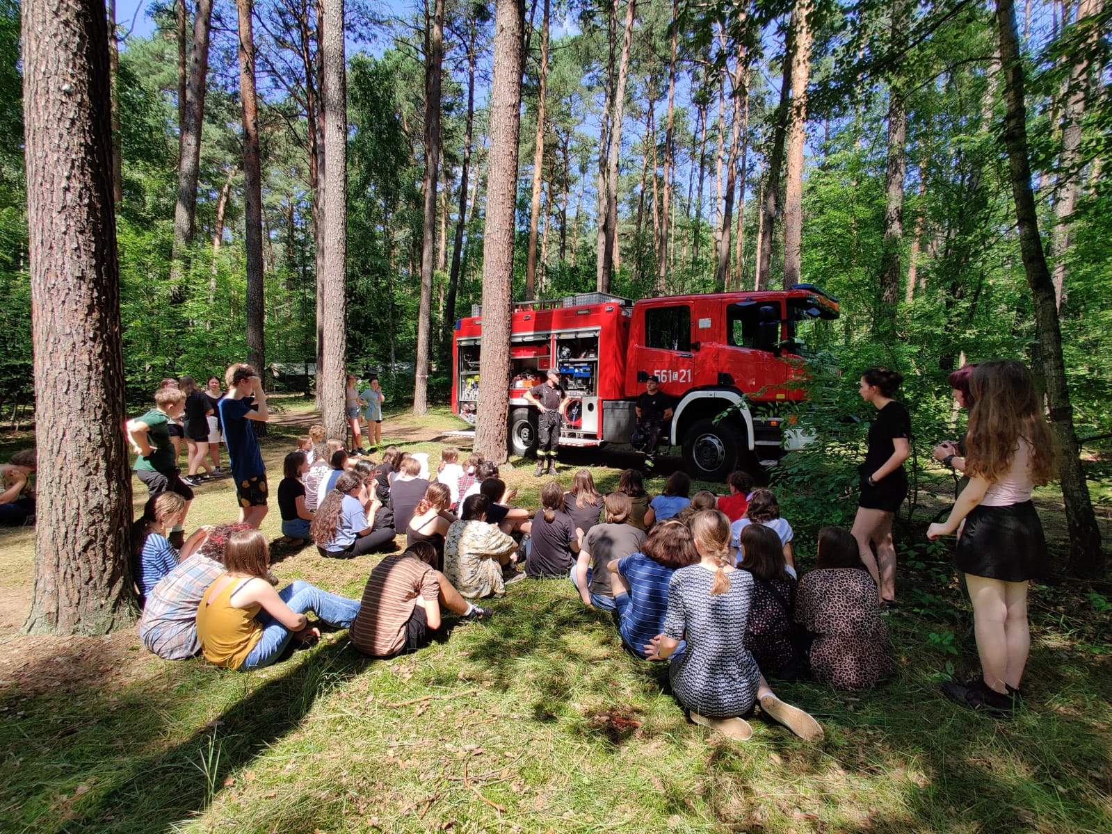W lesie siedzą siedzi, przed nimi strażak stoi przy samochodzie bojowym PSP i pokazuje sprzęt