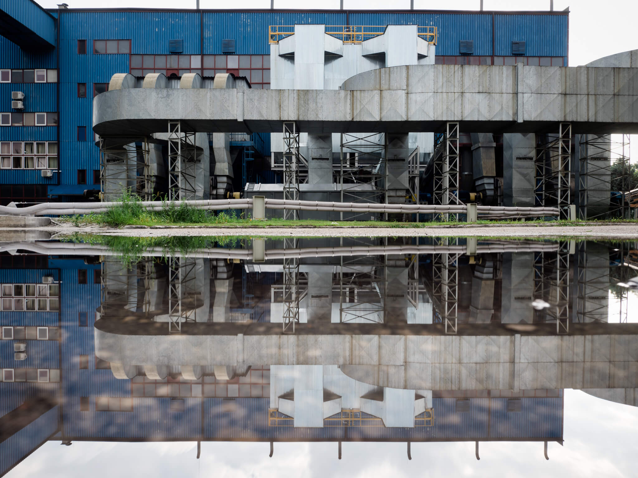 Instalacja ZMIERZ jest dopasowana do istniejącej infrastruktury. Na zdjęciu widoczny pomalowaną na niebiesko halę przemysłową wraz z instalacją w kolorze metalowym oraz jej odbicie w stawie.