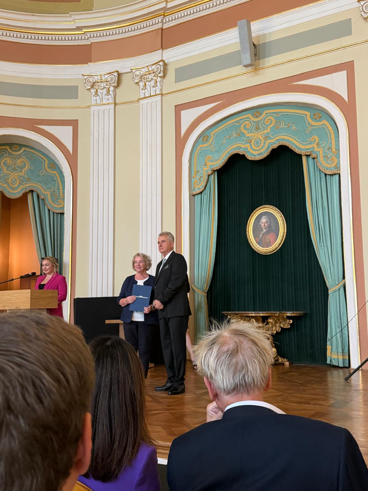 W zabytkowym wnętrzu odbywa się oficjalna ceremonia. Na pierwszym planie widać tyły głów publiczności. Głębiej, w centrum sceny, stoi uśmiechnięta kobieta w granatowym żakiecie, trzymająca niebieską teczkę - Pani Irena Maciejewska. Obok niej stoi mężczyzna w czarnym garniturze - Sławomir Rogowski. Po lewej stronie, za drewnianą mównicą, przemawia kobieta w jaskraworóżowej marynarce. Tło stanowi ozdobna ściana z wnękami zasłoniętymi turkusowymi zasłonami, we wnęce po prawej wisi owalny portret w złotej ramie
