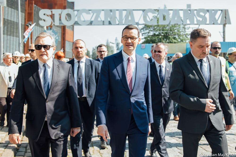 Premier Mateusz Morawiecki (w środku), wicepremier Piotr Gliński i przewodniczący NSZZ Solidarność Piotr Duda idą, a za nimi Stocznia Gdańska.