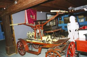 Ręczna sikawka zamocowaną na konnej bryczce. Pojazd znajduje się wewnątrz pomieszczenia, w którym umieszczono inne pożarnicze eksponaty historyczne. 