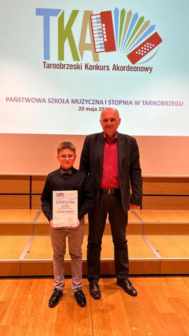 Zdjęcie ukazuje ucznia trzymającego dyplom laureata, a obok niego stoi nauczyciel. Z tyłu widoczny plakat promujący Tarnobrzeski Konkurs Akordeonowy.