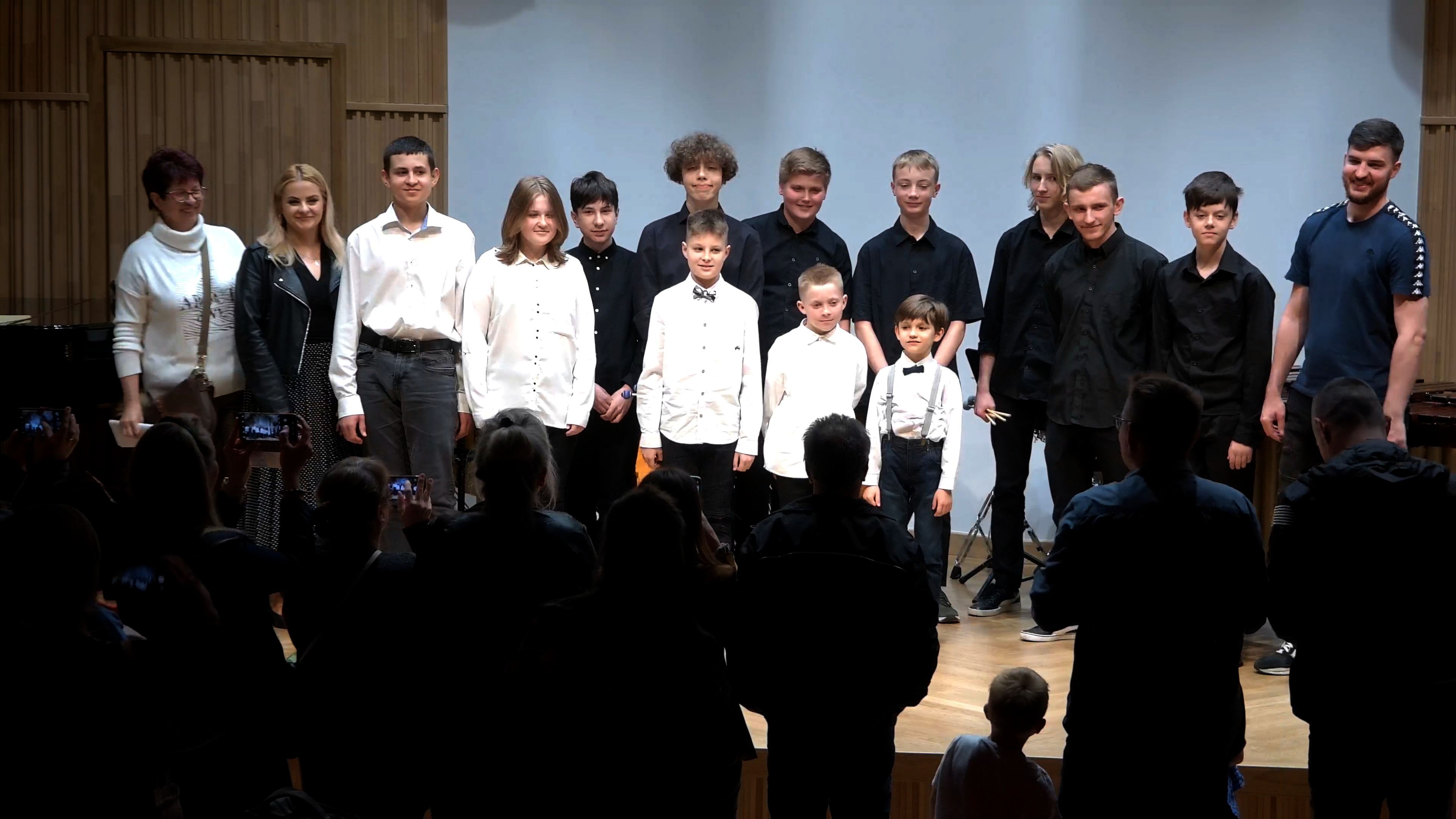 Zdjęcie. Na estradzie - przed publicznością, która stoi i robi zdjęcia - stoją w grupie wykonawcy koncertu i pozują do zdjęcia.