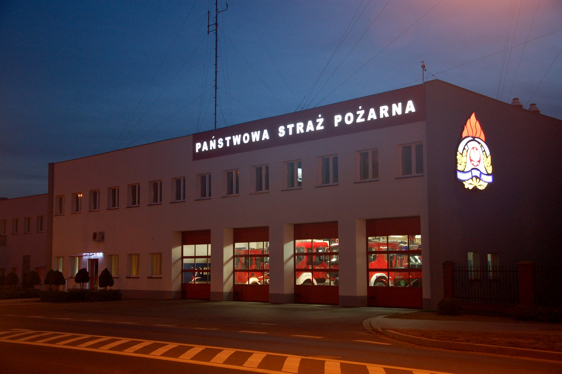 Zdjęcie przedstawia budynek Komendy Powiatowej PSP w Brodnicy od strony ulicy Sienkiewicza po zmierzchu. Na budynku podświetlony napis Państwowa Straż Pożarna oraz logo PSP. W przeszklonych wrotach garażowych widoczny oświetlone pojazdy Jednostki Ratowniczo-Gaśniczej.
