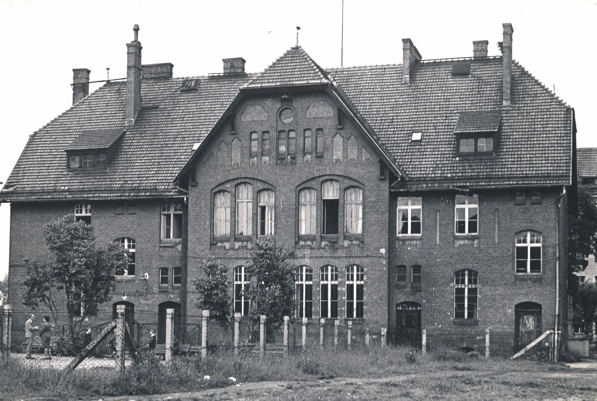 Zdjęcie przedstawia pierwszy budynek szkoły od strony podwórza, które ogrodzone jest płotem. Na podwórzu w dolny lewym rogu ówczesny dyrektor szkoły Tadeusz Rękas rozmawia z jedną z nauczycielek. W pobliżu dwoje małych uczniów. 