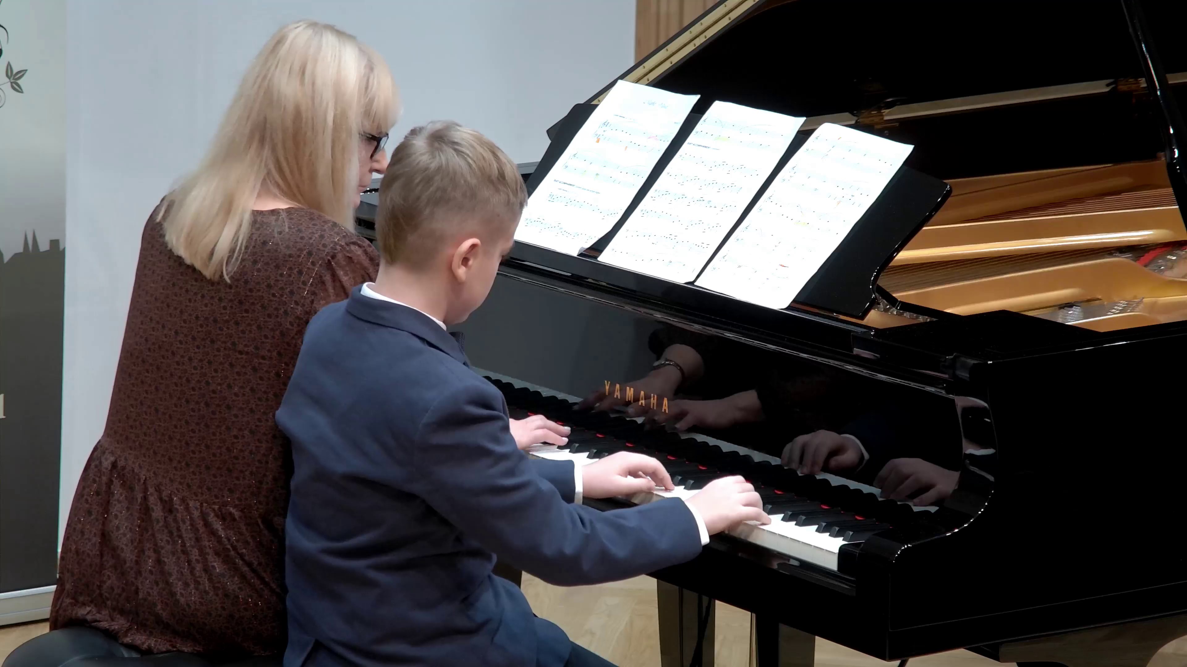 Zdjęcie przedstawia Salę Koncertową PSM w Nysie podczas koncertu uczniów klas fortepianu. W dolnej części słuchacze na widowni, zapowiada pani Bogusława Kos. W głębi estrada, na której stoi otwarty czarny fortepian. Zdjęcie jest częścią albumu zdjęć z koncertu, który odbył się 13 grudnia 2022.