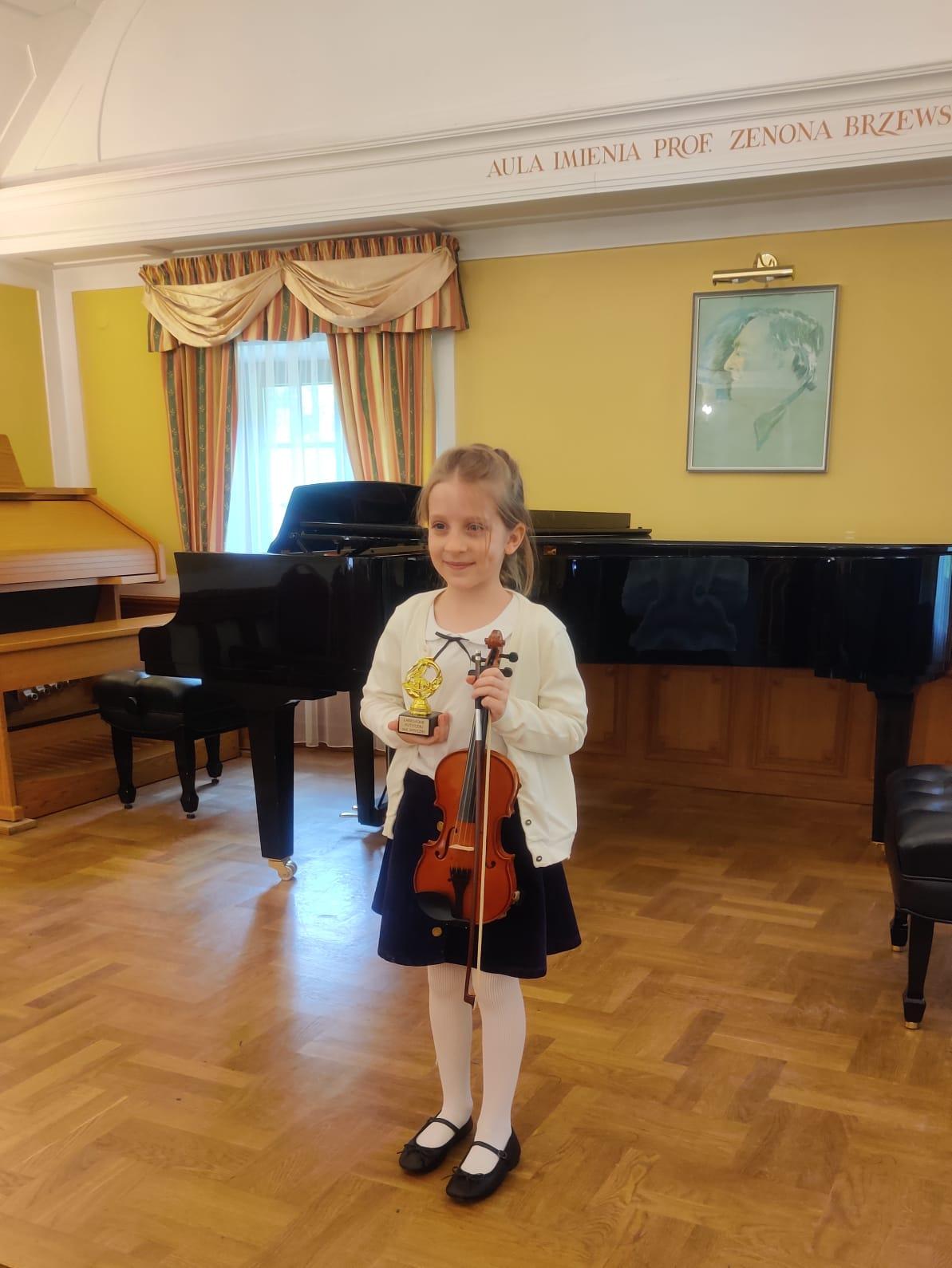 Uczennica trzymająca statuetkę laureata wraz ze skrzypcami. Z tyłu widoczny fortepian i organy.