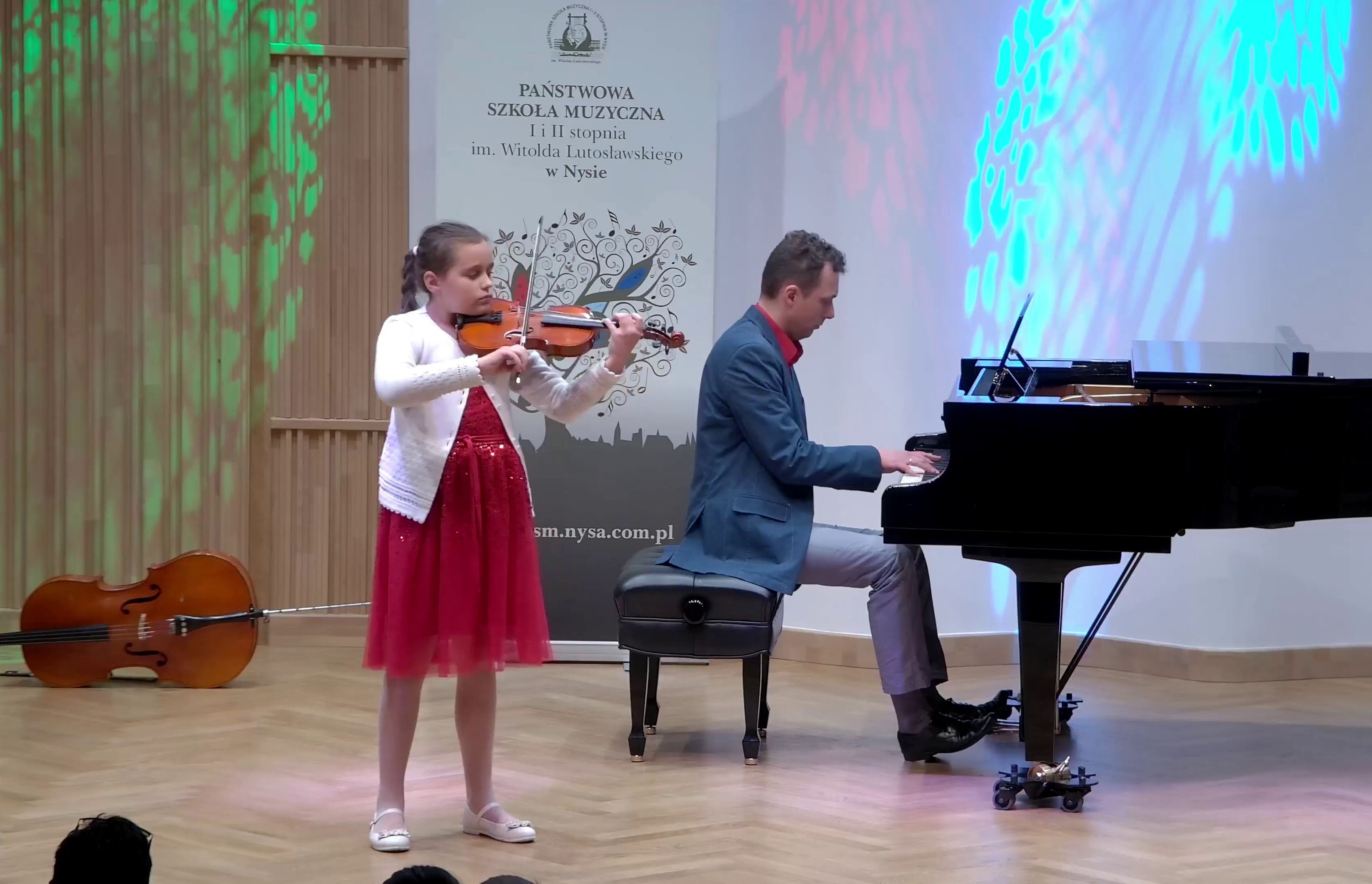 Zdjęcie. Estrada Sali Koncertowej. Występują - młoda skrzypaczka i towarzyszący jej dorosły pianista, grający na dużym czarnym fortepianie. W tle po lewej stronie na podłodze leży wiolonczela, a w środku na białym tle stoi baner PSM w Nysie.