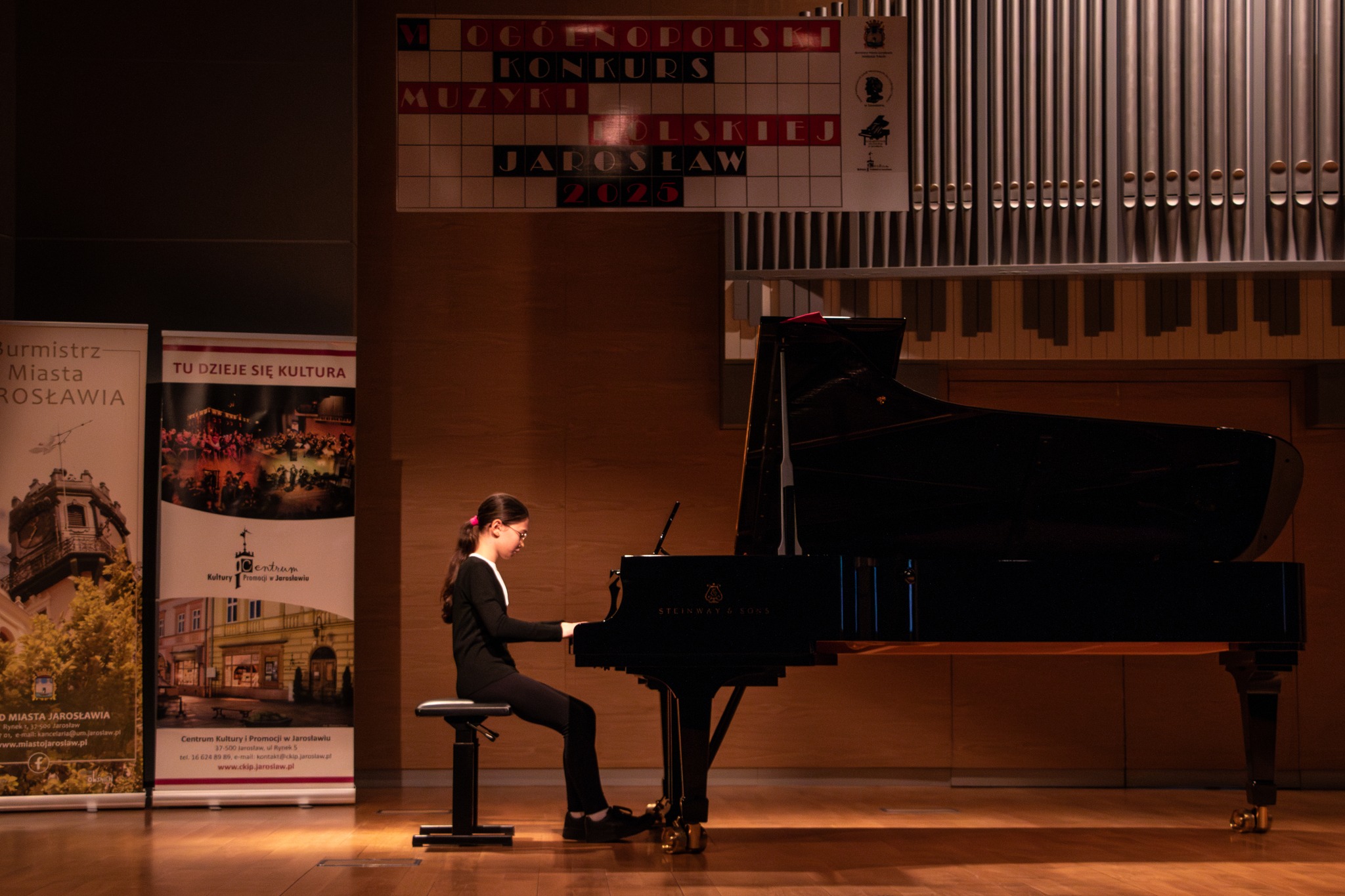 Na drewnianej scenie, oświetlonej z góry, siedzi młoda osoba grająca na czarnym fortepianie. Osoba ta ma ciemne włosy z różową spinką i ubrana jest w ciemną bluzkę z długimi rękawami oraz ciemne spodnie. Siedzi na regulowanym, czarnym stołku przed instrumentem, z rękami ułożonymi na klawiszach. Za fortepianem znajduje się ściana z drewnianą boazerią, a nad nią widoczne są fragmenty organów piszczałkowych. Na ścianie wisi również plakat informujący o "Ogólnopolskim Konkursie Muzyki Polskiej" w Jarosławiu w 2025 roku. Po lewej stronie sceny stoją dwa pionowe banery z tekstami i grafikami w języku polskim, częściowo zasłonięte przez cień. Scena sali koncertowej
