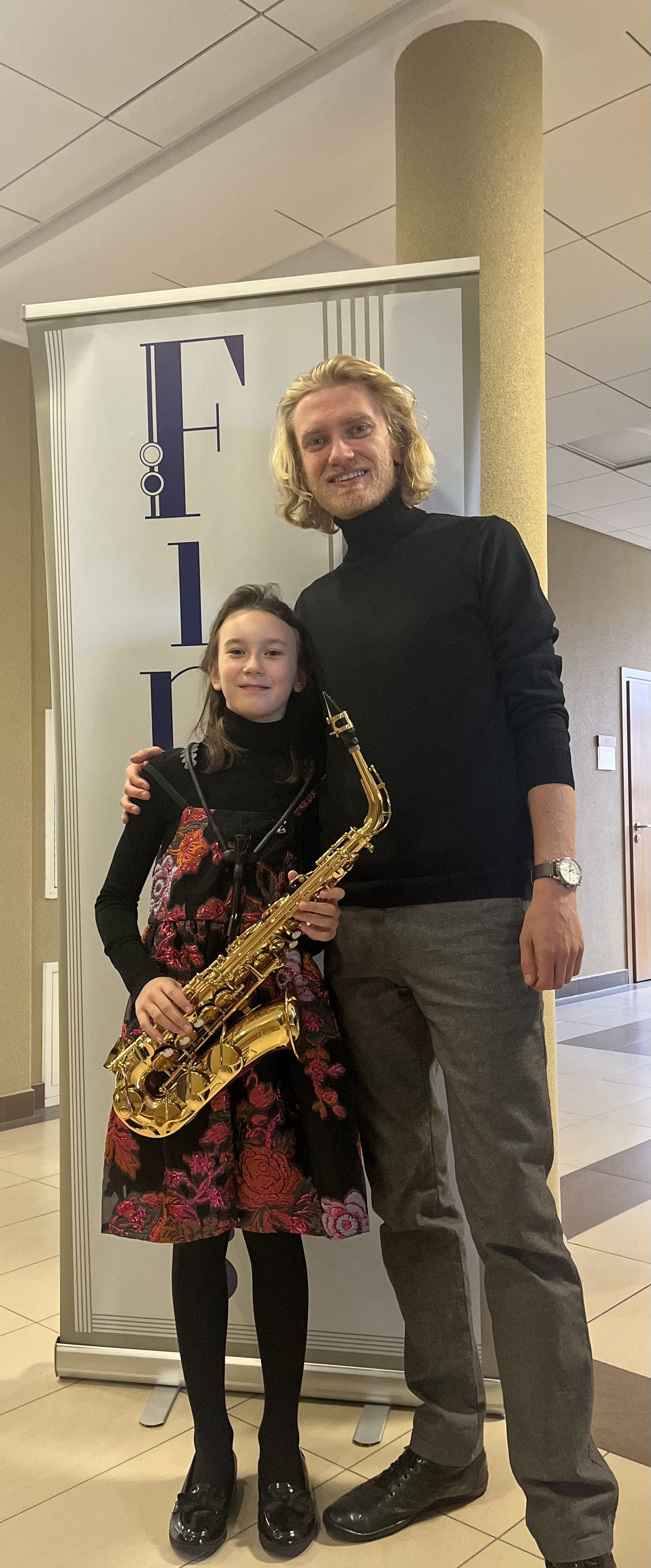 Zdjęcie przedstawia dziewczynkę ubraną w czarny golf i sukienkę w czerwone kwiaty, która trzyma złoty saksofon oraz dorosłego mężczyznę ubranego w czarny golf i szare spodnie. W tle widać baner konkursu Finalis.